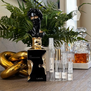 May include: A black and gold perfume bottle with the text "His Confession" and "Lattafa". The bottle is topped with a black bust with gold accents. Several clear sample bottles are next to the perfume bottle, with labels indicating sizes.
