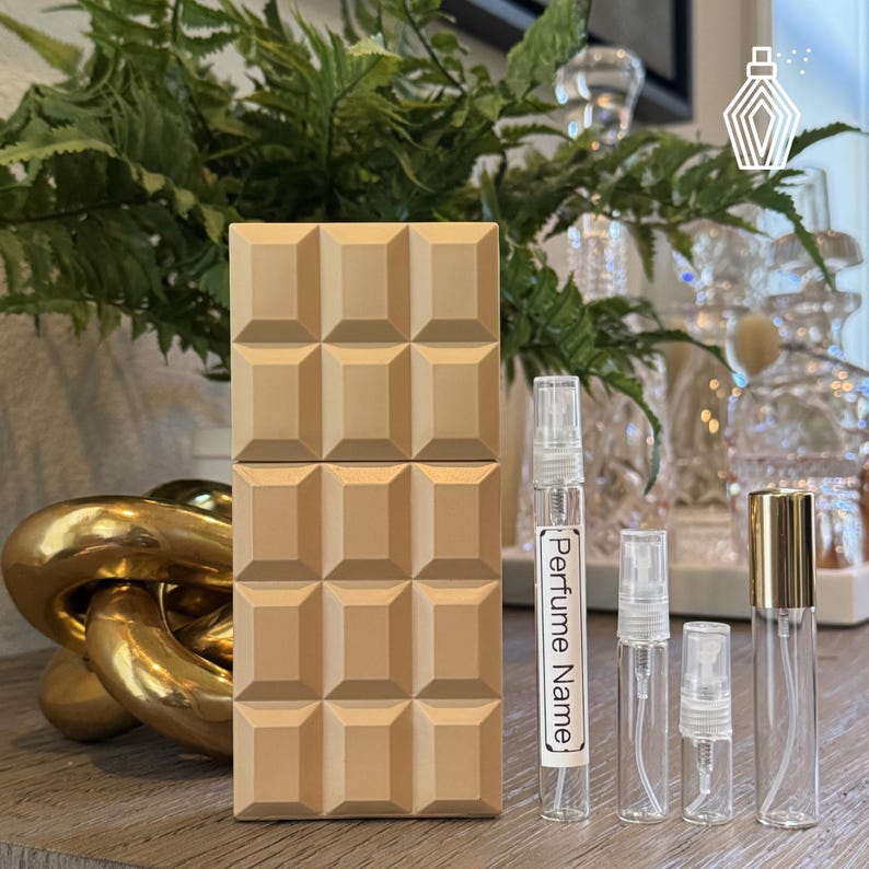 K&ouml;nnte beinhalten: Eine beige Parf&uuml;mflasche mit geometrischem Design, neben mehreren durchsichtigen Glas-Parf&uuml;mfl&auml;schchen. Ein Fl&auml;schchen tr&auml;gt die Aufschrift "Perfume Name". Der Hintergrund umfasst Gr&uuml;n und dekorative Glasflaschen.