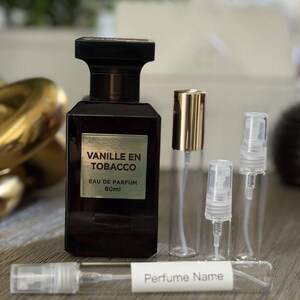 May include: A dark brown perfume bottle with a black cap and a green label that reads "Vanille En Tobacco Eau de Parfum 80ml." Several clear glass vials with sprayers and a vial with a label that reads "Perfume Name" are also present.