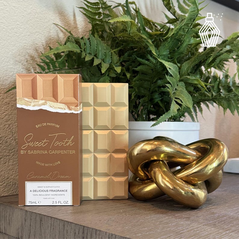K&ouml;nnte beinhalten: Eine Parf&uuml;mbox und ein dekoratives Objekt werden auf einer Holzoberfl&auml;che pr&auml;sentiert. Die Parf&uuml;mbox ist braun und beige, mit dem Text "Sweet Tooth" und "Caramel Dream". Eine goldfarbene Kettenskulptur und ein beiger Block sind ebenfalls vorhanden. Eine Topfpflanze befindet sich im Hintergrund.