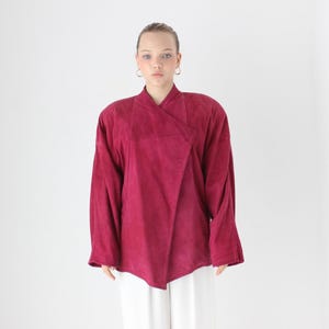 May include: A burgundy wrap-style top with long sleeves. The top has a unique asymmetrical design and a soft, textured appearance. The model is wearing white pants. This top is suitable for various occasions.