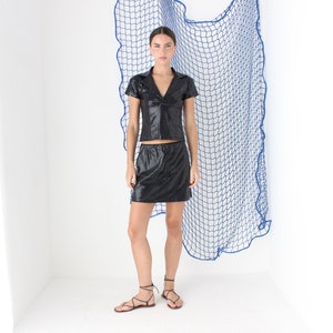 May include: A woman wearing a black leather collared shirt and matching skirt. She is standing in front of a blue net backdrop.