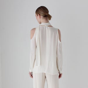Puede incluir: Una blusa blanca de manga larga con un lazo en el cuello y hombros descubiertos.