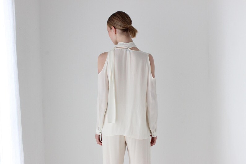 Puede incluir: Una blusa blanca de manga larga con un lazo en el cuello y hombros descubiertos.