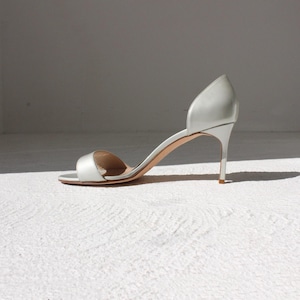 K&ouml;nnte beinhalten: Eine einzelne silberne High-Heel-Sandale mit Stiletto-Absatz. Der offene Schuh hat einen Riemen &uuml;ber den Zehen und ein geschwungenes Design, das den Kn&ouml;chel umschlie&szlig;t. Der Schuh ist vor einem wei&szlig;en Hintergrund platziert.