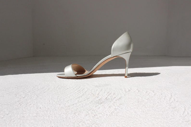 K&ouml;nnte beinhalten: Eine einzelne silberne High-Heel-Sandale mit Stiletto-Absatz. Der offene Schuh hat einen Riemen &uuml;ber den Zehen und ein geschwungenes Design, das den Kn&ouml;chel umschlie&szlig;t. Der Schuh ist vor einem wei&szlig;en Hintergrund platziert.