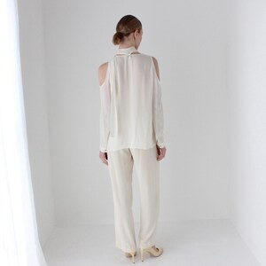 Puede incluir: Una blusa blanca de manga larga con un lazo en el cuello y hombros descubiertos. La blusa se combina con pantalones blancos.