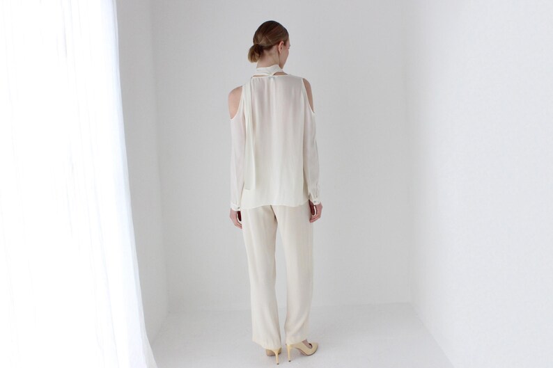 Puede incluir: Una blusa blanca de manga larga con un lazo en el cuello y hombros descubiertos. La blusa se combina con pantalones blancos.