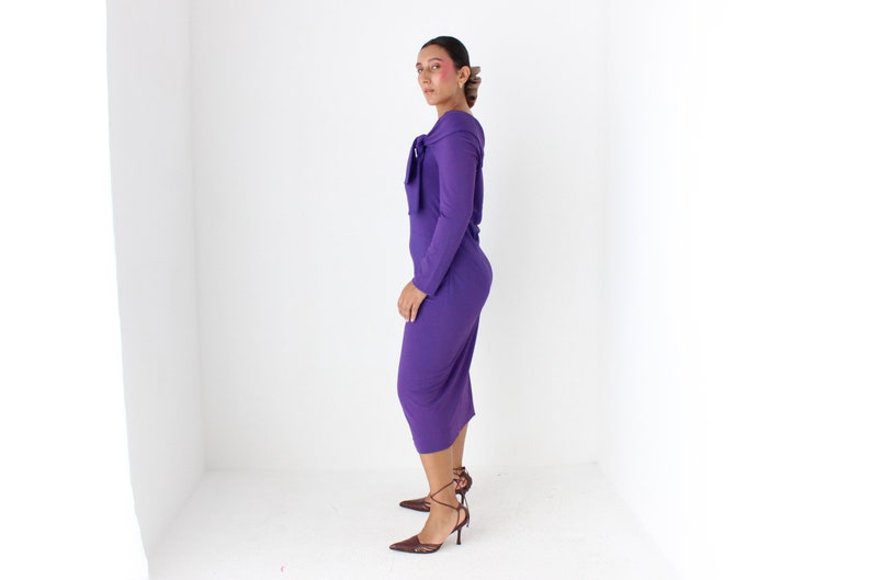 May include: A woman wearing a long-sleeved, purple dress with a tie at the neck. She is standing in a white room and wearing brown heeled shoes.