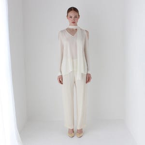 Puede incluir: Una blusa blanca de manga larga con cuello en V y una bufanda blanca atada alrededor del cuello. La blusa tiene hombros descubiertos y la modelo lleva pantalones blancos.