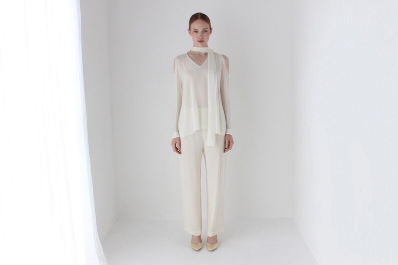 Puede incluir: Una blusa blanca de manga larga con cuello en V y una bufanda blanca atada alrededor del cuello. La blusa tiene hombros descubiertos y la modelo lleva pantalones blancos.