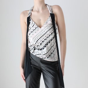 May include: A white and black patterned sleeveless top with a black halter strap and a black fabric draped around the shoulders. The top is paired with black pants.