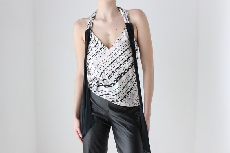 May include: A white and black patterned sleeveless top with a black halter strap and a black fabric draped around the shoulders. The top is paired with black pants.