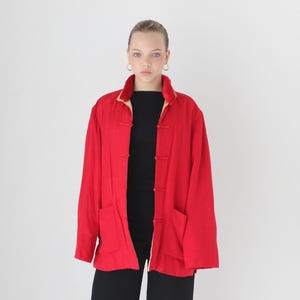 May include: A bright red jacket with a mandarin collar and toggle closures. The jacket has long sleeves and two front pockets. The model is wearing a black top and black pants. The jacket has a yellow lining.