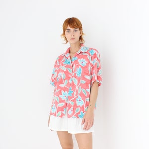 May include: A short-sleeved, button-down shirt in a coral color, adorned with a floral pattern in shades of blue and green. The shirt has a classic collar and is paired with a white skirt. The overall style is casual and summery.