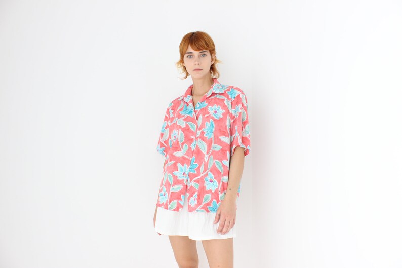 May include: A short-sleeved, button-down shirt in a coral color, adorned with a floral pattern in shades of blue and green. The shirt has a classic collar and is paired with a white skirt. The overall style is casual and summery.