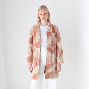 Puede incluir: Un blazer largo de patchwork en tonos melocotón y beige. El blazer tiene un cuello clásico, cierre de botones y mangas largas. Se lleva sobre una camiseta blanca y combinado con pantalones blancos.