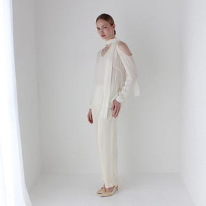 Puede incluir: Una blusa blanca de hombros descubiertos con una larga bufanda fluida atada alrededor del cuello. La blusa tiene un corte holgado y est&aacute; hecha de una tela ligera. La modelo lleva pantalones blancos y tacones beige.