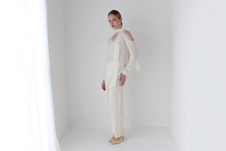 Puede incluir: Una blusa blanca de hombros descubiertos con una larga bufanda fluida atada alrededor del cuello. La blusa tiene un corte holgado y est&aacute; hecha de una tela ligera. La modelo lleva pantalones blancos y tacones beige.