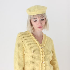 May include: A yellow crocheted cardigan with a scalloped trim and button closure. The cardigan is worn over a white shirt and paired with a yellow crocheted beret.