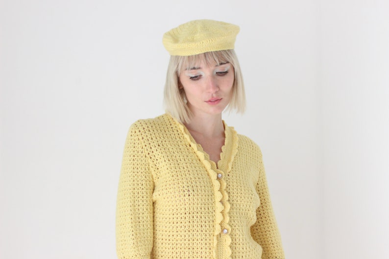 May include: A yellow crocheted cardigan with a scalloped trim and button closure. The cardigan is worn over a white shirt and paired with a yellow crocheted beret.