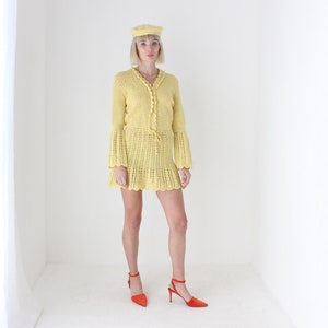 May include: A yellow crochet dress with a crocheted bell sleeve and a matching yellow beret. The dress has a tie at the waist and a scalloped hem. The model is wearing red heeled shoes.