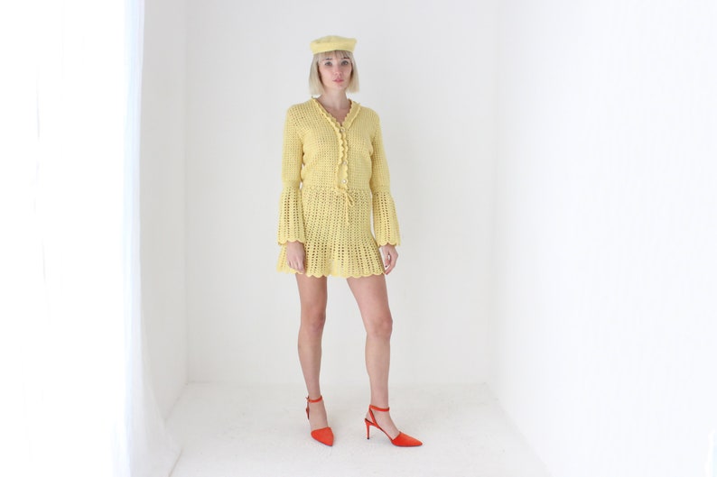 May include: A yellow crochet dress with a crocheted bell sleeve and a matching yellow beret. The dress has a tie at the waist and a scalloped hem. The model is wearing red heeled shoes.