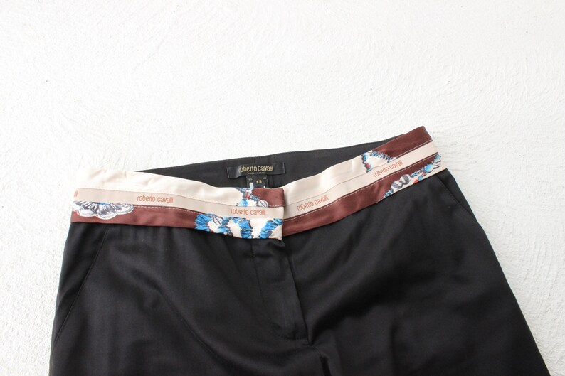 May include: Black pants with a brown and cream patterned waistband. The waistband has the text "Roberto Cavalli" repeated three times.