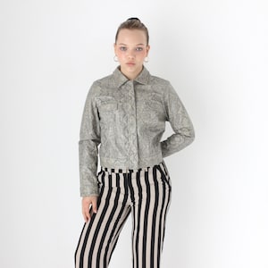 Op de afbeelding: Een zilver-grijze jas met een textuurpatroon, knoopsluiting en twee borstzakken. Het model draagt een broek met zwart-witte strepen. De jas heeft lange mouwen en een kraag. De outfit staat tegen een witte achtergrond.