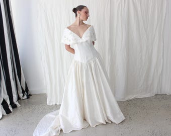 Vestido de novia estilo años 80, de seda cruda dupión color marfil, corte vasco, hombros descubiertos, bordado de perlas y ribete festoneado.