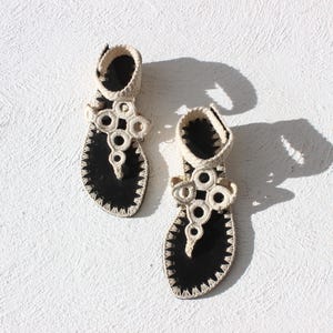 Op de afbeelding: Een paar handgemaakte zwarte sandalen met off-white haakdetails. De sandalen hebben een teenslipper-ontwerp met een decoratief haakpatroon op de bandjes. De zolen zijn zwart met een gestikte rand, tegen een witte achtergrond.