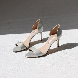 K&ouml;nnte beinhalten: Ein Paar hellgraue High-Heel-Sandalen mit einem schmalen Kn&ouml;chelriemen und einem seitlichen Ausschnitt. Die Abs&auml;tze sind etwa 7,6 cm hoch. Die Schuhe werden auf einer wei&szlig;en Oberfl&auml;che mit Schatten dargestellt.