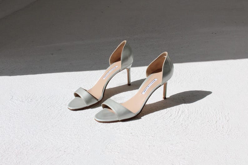 K&ouml;nnte beinhalten: Ein Paar hellgraue High-Heel-Sandalen mit einem schmalen Kn&ouml;chelriemen und einem seitlichen Ausschnitt. Die Abs&auml;tze sind etwa 7,6 cm hoch. Die Schuhe werden auf einer wei&szlig;en Oberfl&auml;che mit Schatten dargestellt.