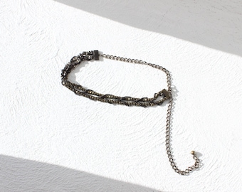 May include: A dark gray choker necklace with a layered design. The necklace features a series of small, sparkling embellishments and a delicate chain extension. The jewelry is set against a white background.