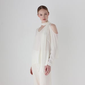 Puede incluir: Una blusa blanca de hombros descubiertos con manga larga y un detalle anudado en el cuello.