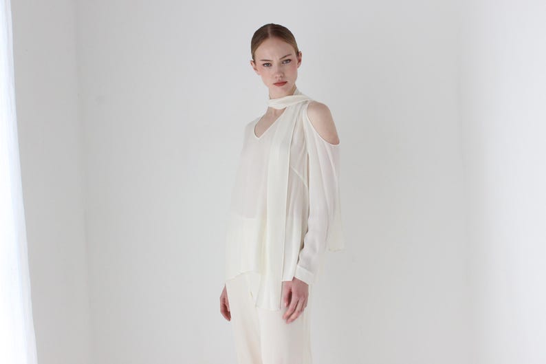 Puede incluir: Una blusa blanca de hombros descubiertos con manga larga y un detalle anudado en el cuello.