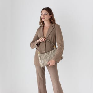 Op de afbeelding: Een bruin damespak met bijpassende handtas. Het pak heeft een blazer en broek met een subtiel krijtstreepmotief. De handtas heeft een slangenprint en een zwarte band.
