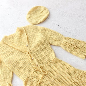May include: A yellow crocheted dress with long sleeves and a matching hat. The dress has a V-neckline and a tie at the waist.