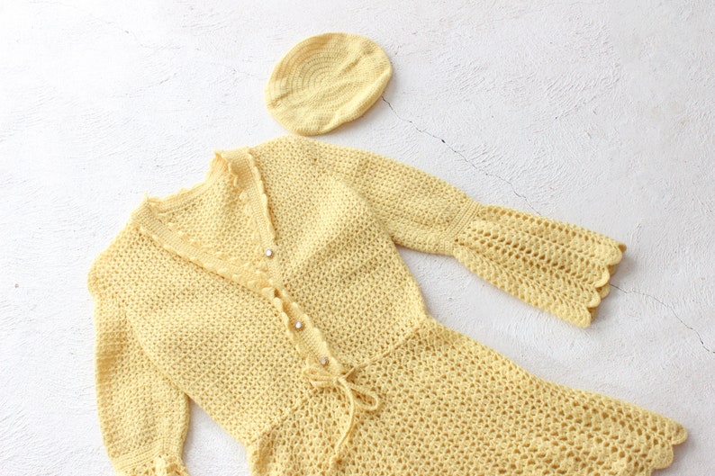 May include: A yellow crocheted dress with long sleeves and a matching hat. The dress has a V-neckline and a tie at the waist.