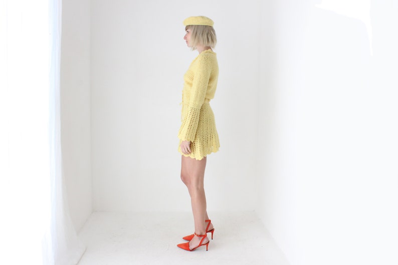 May include: A yellow crocheted dress with a matching beret. The dress has a fitted bodice and a flared skirt. The model is wearing red heels.