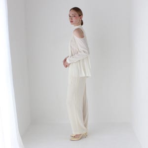 Puede incluir: Una blusa blanca de hombros descubiertos con mangas largas y una silueta fluida. La blusa se combina con pantalones blancos a juego.