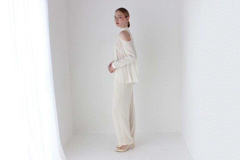 Puede incluir: Una blusa blanca de hombros descubiertos con mangas largas y una silueta fluida. La blusa se combina con pantalones blancos a juego.