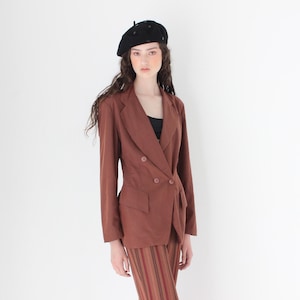 Op de afbeelding: Een roestkleurige double-breasted blazer en bijpassende rok. De blazer heeft twee rijen knopen en een getailleerde pasvorm. De rok heeft verticale strepen in bruin- en roodtinten. Een zwarte baret maakt de outfit compleet.