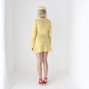 May include: A yellow crochet dress with a ruffled hem and long sleeves. The dress is worn by a person with blonde hair and a yellow hat.