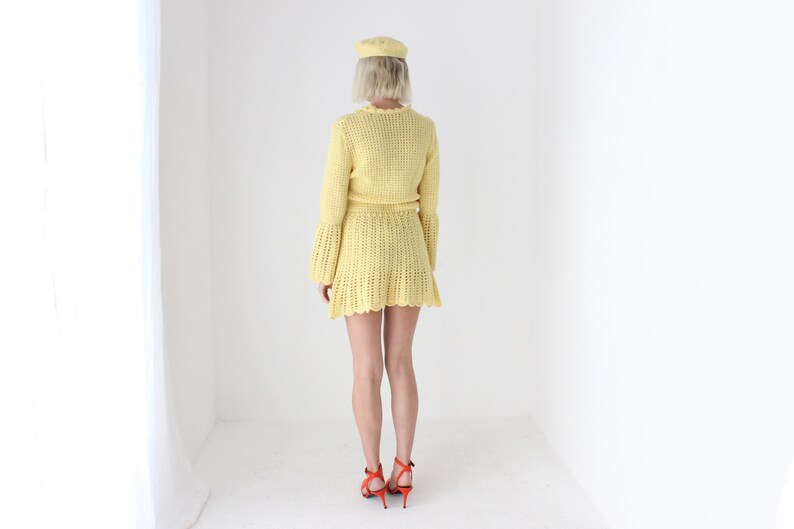 May include: A yellow crochet dress with a ruffled hem and long sleeves. The dress is worn by a person with blonde hair and a yellow hat.