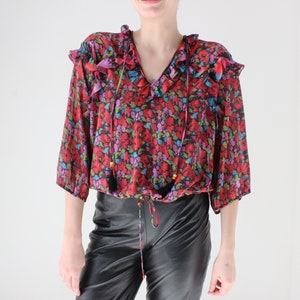 May include: A red, blue, and green floral print blouse with a ruffled neckline and tassel ties. The blouse has a gathered waist and 3/4 sleeves.