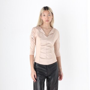 May include: A beige-colored blouse with a V-neck and three-quarter sleeves. The blouse has a tailored fit with vertical seams down the front. The sleeves have button details. The model is wearing black leather pants.