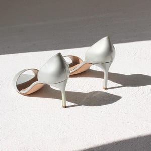 K&ouml;nnte beinhalten: Ein Paar silberne High-Heel-Sandalen mit spitzer Zehenpartie. Die Schuhe haben Stiletto-Abs&auml;tze und sind aus gl&auml;nzendem Material. Sie liegen auf einer wei&szlig;en Oberfl&auml;che, mit Schatten.