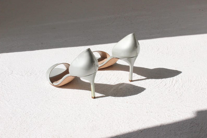 K&ouml;nnte beinhalten: Ein Paar silberne High-Heel-Sandalen mit spitzer Zehenpartie. Die Schuhe haben Stiletto-Abs&auml;tze und sind aus gl&auml;nzendem Material. Sie liegen auf einer wei&szlig;en Oberfl&auml;che, mit Schatten.