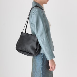 May include: Black leather shoulder bag with a curved bottom and long, thin straps. The bag is worn over the shoulder. The bag is a solid black color and appears to be made of leather. The background is a plain white.